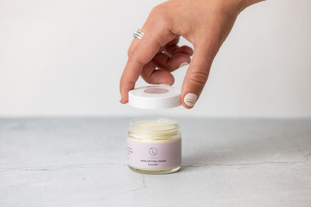 Hand opening a jar of CBD Lavender Shea Butter cream, showcasing the hydrating body lotion inside.