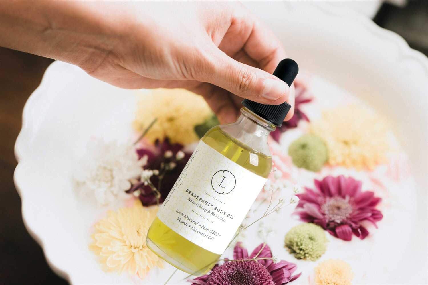 Hand holding a bottle of grapefruit moisturizing body oil surrounded by colorful flowers.