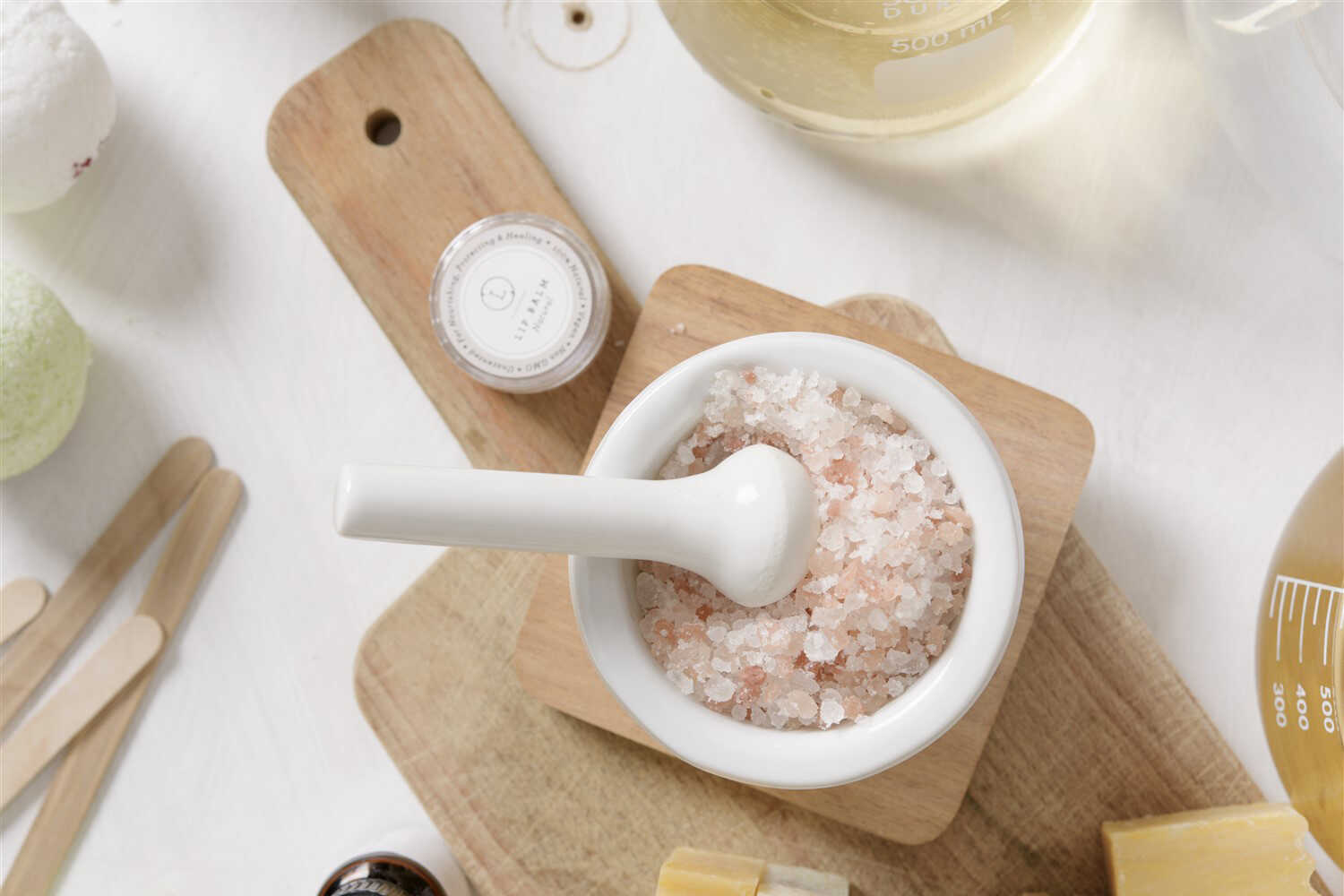 White mortar and pestle with pink bath salts on a wooden board, surrounded by spa items.